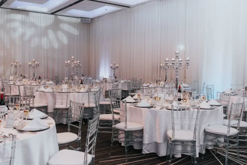 The image shows an elegant wedding venue at Plaza Centre Ville, categorized under Wedding Venues. The spacious room features round tables adorned with white tablecloths and neatly folded napkins. Each table is elegantly set with fine china, glassware, and silverware. Tall, ornate candelabras serve as centerpieces, enhancing the venue's sophisticated ambiance. The room is softly illuminated with intricate light patterns projected onto the white draped walls, creating a romantic and inviting atmosphere perfect for hosting an elegant wedding reception.