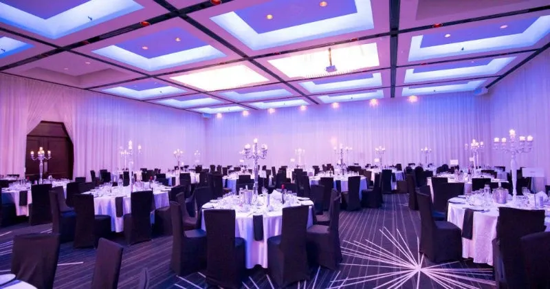 The image showcases an elegantly arranged wedding venue at Plaza Centre Ville, categorized under Wedding Venues on Mariage Elegant, Canada. The spacious room features multiple round tables adorned with crisp white tablecloths, each set with fine china and glassware, ready for a wedding reception. Tall, sophisticated candelabras serve as centerpieces, adding to the ambiance. The ceiling is illuminated with soft, ambient lighting that casts a warm glow, enhancing the romantic atmosphere. The venue's modern decor and ample space make it an ideal setting for a sophisticated wedding celebration.