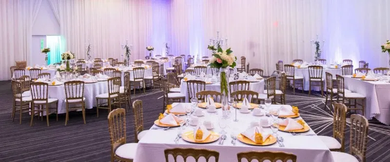 This image showcases the elegant interior of Plaza Centre Ville, a prominent wedding venue in Canada listed on Mariage Elegant. The spacious hall is adorned with round tables draped in pristine white tablecloths, each elegantly set with fine china and folded yellow napkins. Gold chiavari chairs surround the tables, adding a touch of sophistication. Tall floral centerpieces and candelabras with soft candlelight create a romantic ambiance. The backdrop features sheer white drapes with mood lighting in soft purple hues, enhancing the venue's luxurious atmosphere, perfect for hosting an unforgettable wedding celebration.