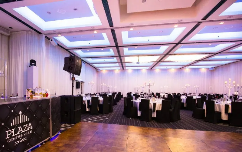 The image features the Plaza Centre Ville, a premier wedding venue in Canada, elegantly set up for a wedding reception. The spacious room is adorned with white draped walls and a beautifully lit ceiling featuring recessed lighting panels. Numerous round tables are arranged neatly across the venue, each elegantly dressed with white tablecloths and black chair covers. The tables are adorned with intricate candelabras, adding a touch of sophistication and ambiance. A polished wooden dance floor is visible in the foreground, adjacent to a chic bar area featuring the venue's logo. This stunning setup highlights the Plaza Centre Ville's capability to host upscale wedding events.