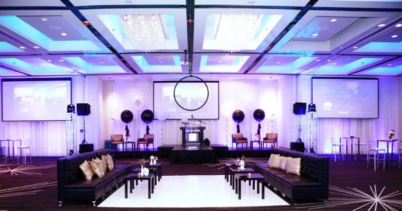 The image showcases a luxurious wedding venue at Plaza Centre Ville, categorized under wedding venues on Mariage Elegant, Canada. The spacious hall is elegantly decorated with modern features, including a polished white dance floor, surrounded by black sofas with gold accent pillows. Overhead, the ceiling is adorned with sparkling chandeliers, casting a warm glow throughout the room. Large projection screens are visible on the walls, suggesting advanced audio-visual capabilities for wedding presentations or entertainment. The overall ambiance conveys sophistication and readiness to host a stylish wedding event.