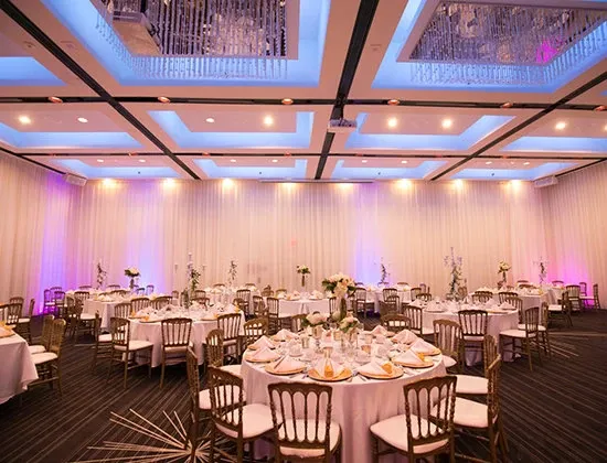 The image showcases an elegant wedding venue, Plaza Centre Ville, under the wedding venues category. The spacious room features tastefully arranged round tables adorned with white tablecloths and beautifully set with fine china and glassware. Each table is complemented with floral centerpieces adding a touch of romance. The ceiling is embellished with modern, recessed lighting and decorative panels, casting a soft, ambient glow. The venue's draped walls and sophisticated decor create a luxurious atmosphere, perfect for an elegant wedding reception.