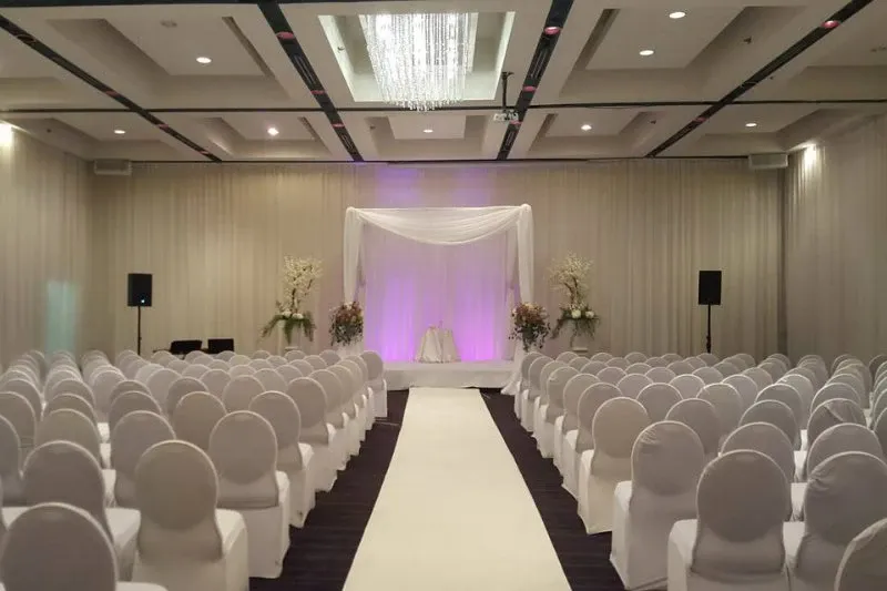 The image shows an elegantly decorated wedding venue at Plaza Centre Ville, a listed vendor in the wedding category on Mariage Elegant, Canada. The spacious room features rows of neatly arranged white chairs lining both sides of a central aisle covered with a white runner. At the end of the aisle, there is a beautifully draped altar with a soft white fabric canopy, enhanced by subtle purple lighting in the background. The atmosphere is sophisticated and serene, suitable for a wedding ceremony. Floral arrangements on pedestals flank the altar, adding a touch of natural beauty to the setting.