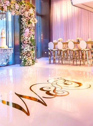 The image showcases a beautifully decorated wedding venue at Plaza Centre Ville, highlighting its suitability for elegant wedding celebrations. The scene features a luxurious hall with a glossy white floor adorned with intricate gold swirling designs. To the left, there's a tall, cascading floral arrangement with soft pink and white flowers that adds a romantic touch. In the background, round tables with gold frames and white cushioned chairs are neatly arranged, set against a backdrop of sheer pink drapery that creates a warm and inviting ambiance. The setting at this wedding venue perfectly combines sophistication and charm, ideal for hosting memorable weddings in Canada.