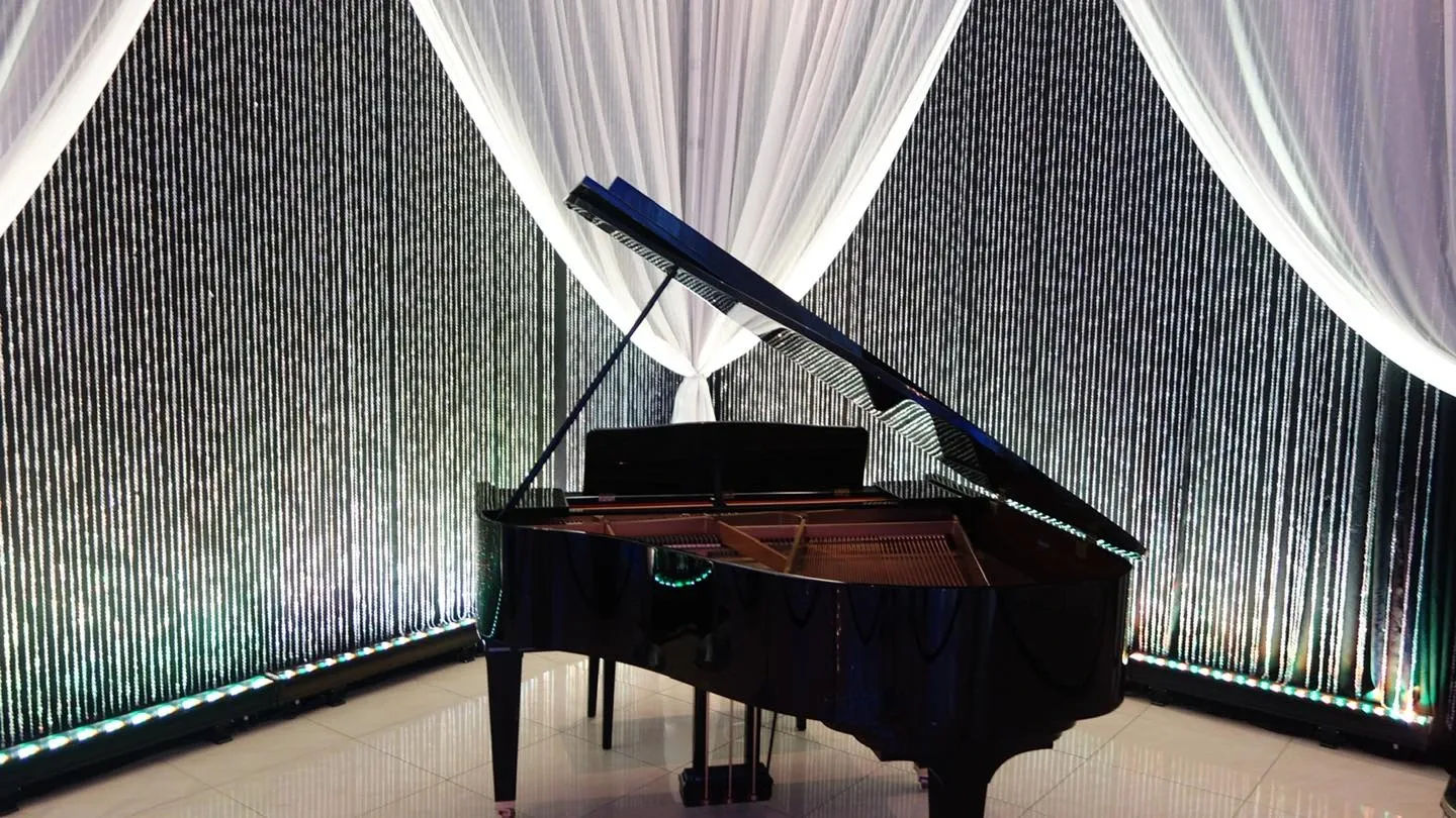 The image showcases a grand piano set at the Embassy Plaza, a distinguished venue in the wedding category. The piano is placed elegantly in the corner of a room adorned with cascading string curtains and flowing white drapes, creating a sophisticated and romantic ambiance. The setting is well-lit, emphasizing a luxurious and stylish atmosphere ideal for wedding ceremonies or receptions.