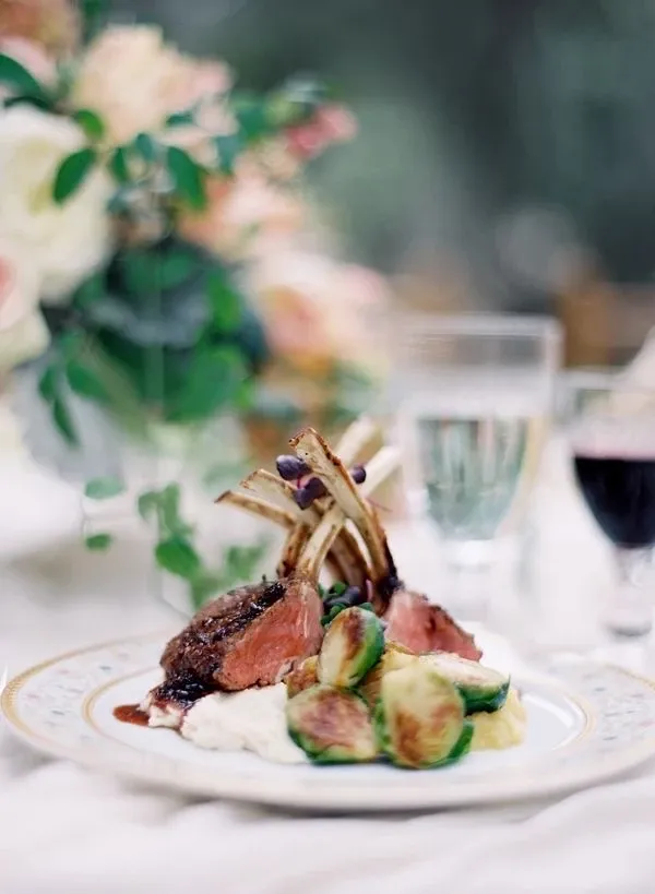 The image showcases an elegant dining setting at Embassy Plaza, a premium wedding venue in Canada. In the foreground, a beautifully plated gourmet dish features succulent lamb chops elegantly arranged over a bed of creamy mashed potatoes and garnished with Brussels sprouts. The table is adorned with a delicate floral arrangement in soft pastel hues, enhancing the romantic ambiance. In the background, crystal-clear wine glasses are filled with white and red wine, adding a touch of sophistication to the exquisite wedding dining experience crafted by this venue.