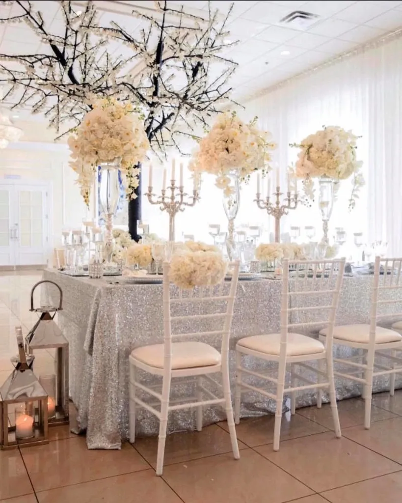 This image showcases a beautifully decorated wedding venue at Embassy Plaza, categorized under Wedding Venues on Mariage Elegant, Canada. The setup features an elegant, long table draped with a shimmering silver tablecloth, adorned with tall, clear vases filled with lush white flower arrangements. The arrangement is complemented by ornate silver candelabras, adding a touch of sophistication. The chairs are chic and white, fitting the luxurious theme. Overhead, branches with delicate white blossoms are artistically displayed, enhancing the romantic ambiance. To the side, lanterns with candles add warmth and charm to the setting. The bright, airy room with sheer curtains completes this exquisite wedding scene.