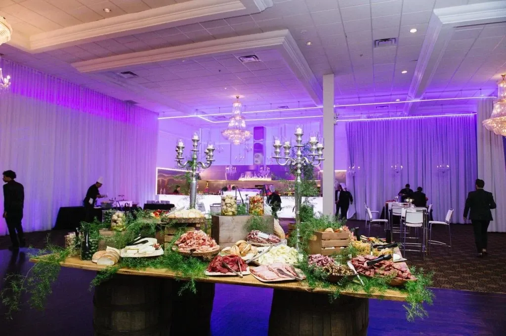 A beautifully decorated wedding reception hall at Embassy Plaza, showcasing a lavish grazing table with an assortment of cured meats, cheeses, and fresh bread, elegantly arranged on wooden barrels. The venue, classified under the Wedding Venues category, features sophisticated chandeliers and soft purple uplighting that complement the draped white curtains surrounding the room. Several guests and staff are visible, enhancing the lively atmosphere. This setting is ideal for couples seeking an elegant and memorable celebration.