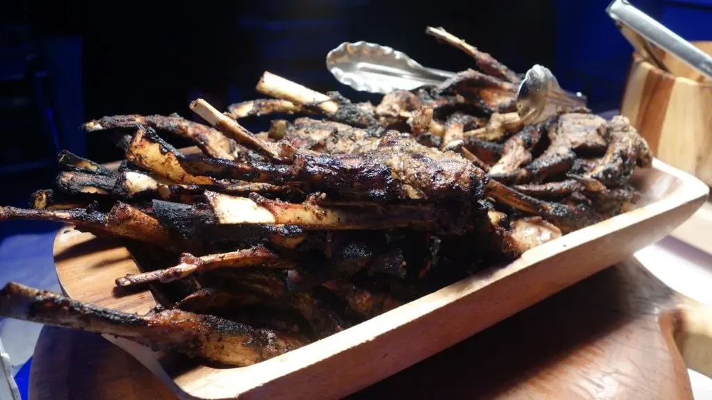 This image showcases a wooden platter filled with grilled lamb chops, presented as part of the culinary offerings at Embassy Plaza, a renowned wedding venue featured on Mariage Elegant. The lamb chops are expertly charred, highlighting their rich, savory flavor, making them an ideal choice for wedding receptions. The platter is accompanied by a pair of metal tongs, suggesting a buffet-style service commonly seen in elegant wedding settings. The focus on high-quality catering underscores Embassy Plaza's reputation for providing exceptional dining experiences for weddings and special events in Canada.