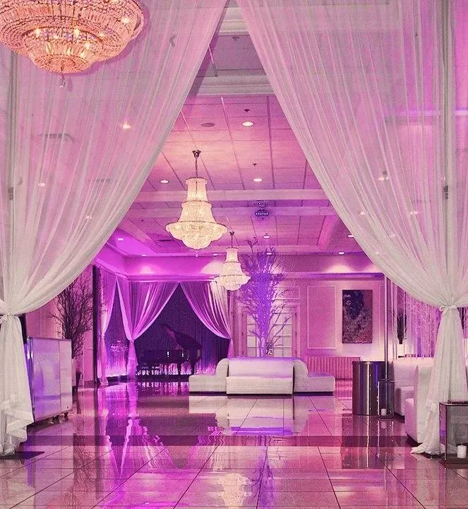 The image shows the interior of Embassy Plaza, a wedding venue listed under Mariage Elegant in Canada. The elegant setting features a grand entrance framed by sheer white curtains, leading to a spacious hall bathed in soft pink and purple lighting. Sparkling chandeliers hang from the ceiling, reflecting light across the polished tiled floor. A stylish lounge area with a white sofa is positioned towards the back, offering a cozy space for guests. The decor includes tall decorative branches and sophisticated wall art, enhancing the sophisticated ambiance ideal for a wedding celebration.