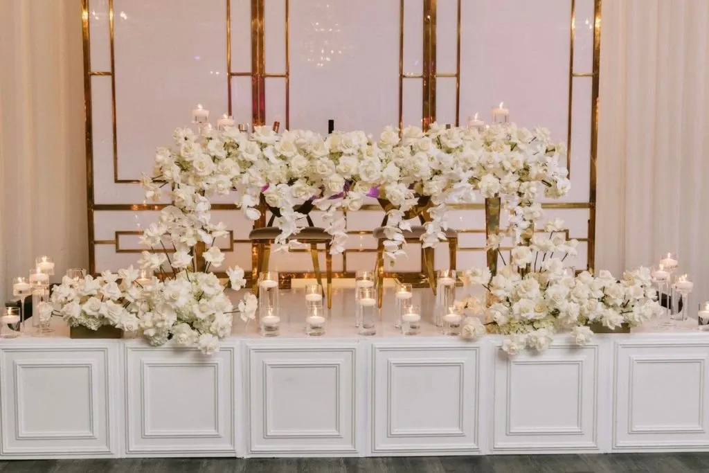 The image showcases a beautifully decorated wedding venue at Embassy Plaza, listed under the Wedding Venues category on Mariage Elegant, Canada. A stunning altar is adorned with an abundance of white roses and elegant greenery. The floral arrangements are complemented by numerous candles in glass holders, creating a romantic and warm ambiance. The backdrop features a sophisticated geometric design with gold accents, enhancing the luxurious feel of the venue. The setting is perfectly tailored for an elegant wedding ceremony, providing a picturesque and enchanting environment.