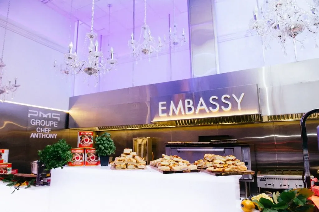 The image showcases the spacious and elegantly decorated interior of Embassy Plaza, a top-rated wedding venue listed on Mariage Elegant in Canada. The room is adorned with modern crystal chandeliers hanging from the ceiling, casting a soft glow over the venue. In the foreground, there are beautifully arranged trays of gourmet sandwiches, suggesting a catering setup. The prominent Embassy Plaza signage is visible on the sleek, reflective wall, highlighting the venue's stylish atmosphere. This image effectively captures the sophisticated environment suitable for hosting weddings and special events.