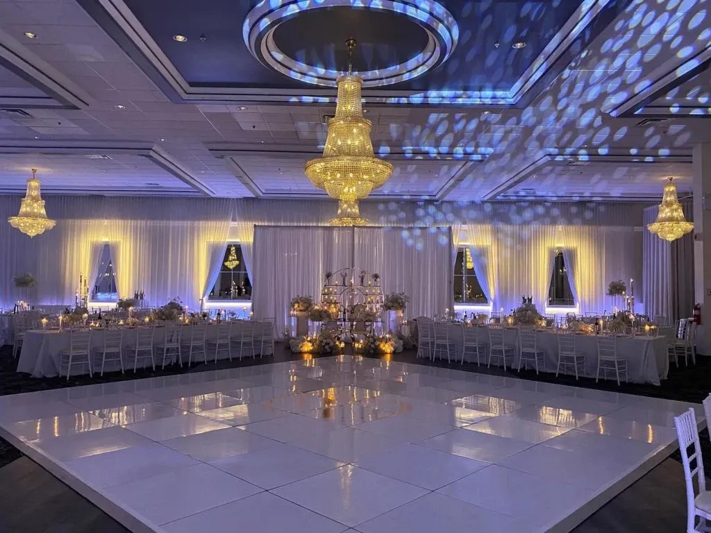 The image shows a beautifully decorated wedding venue at Embassy Plaza, categorized under Wedding Venues on Mariage Elegant, Canada. The spacious hall features elegant chandeliers and ambient blue and yellow lighting that sets a romantic atmosphere. A large, polished white dance floor is centered, surrounded by tables adorned with white linens and floral centerpieces. Chiavari chairs complement the sophisticated decor, enhancing the venue's luxurious and welcoming ambiance.