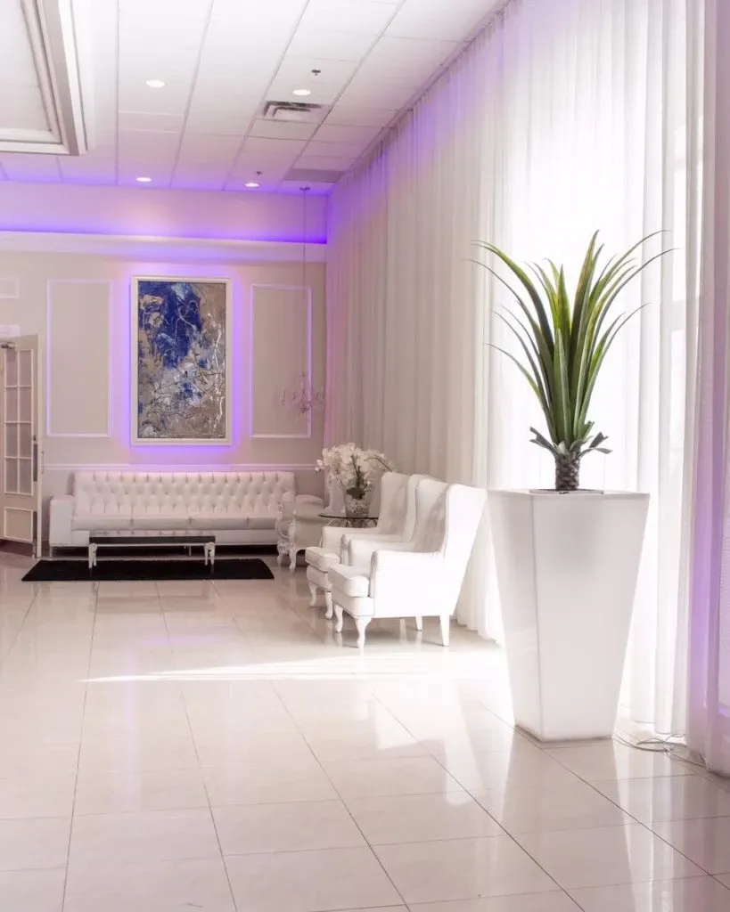 The image features a sophisticated interior of Embassy Plaza, a wedding venue in Canada, showcasing an elegantly designed lounge area. The room is adorned with luxurious white sofas and armchairs, complemented by a white coffee table. An abstract painting with blue hues is mounted on the wall, drawing attention and adding a touch of modern art. A large potted plant in a sleek, white vase stands to the right, contributing to the refined decor. Soft purple lighting and sheer white curtains enhance the inviting and romantic ambiance, perfect for weddings and special events.