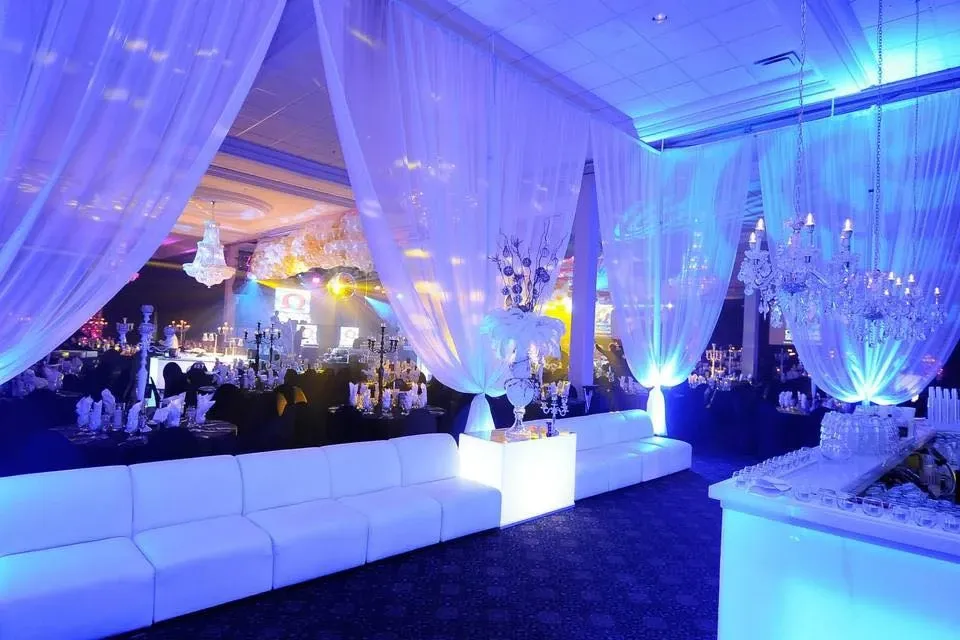 The image showcases a beautifully decorated wedding venue at Embassy Plaza, categorized under wedding venues on Mariage Elegant, Canada. The scene is set with elegant white drapes cascading from the ceiling, illuminated by soft blue and purple lighting that creates a serene and romantic atmosphere. A row of plush white sofas lines the foreground, offering comfortable seating for guests. In the background, exquisite chandeliers add a touch of luxury, hanging above tables adorned with intricate centerpieces. The tables are set with elegant white dinnerware and glasses, ready for a sophisticated wedding reception.
