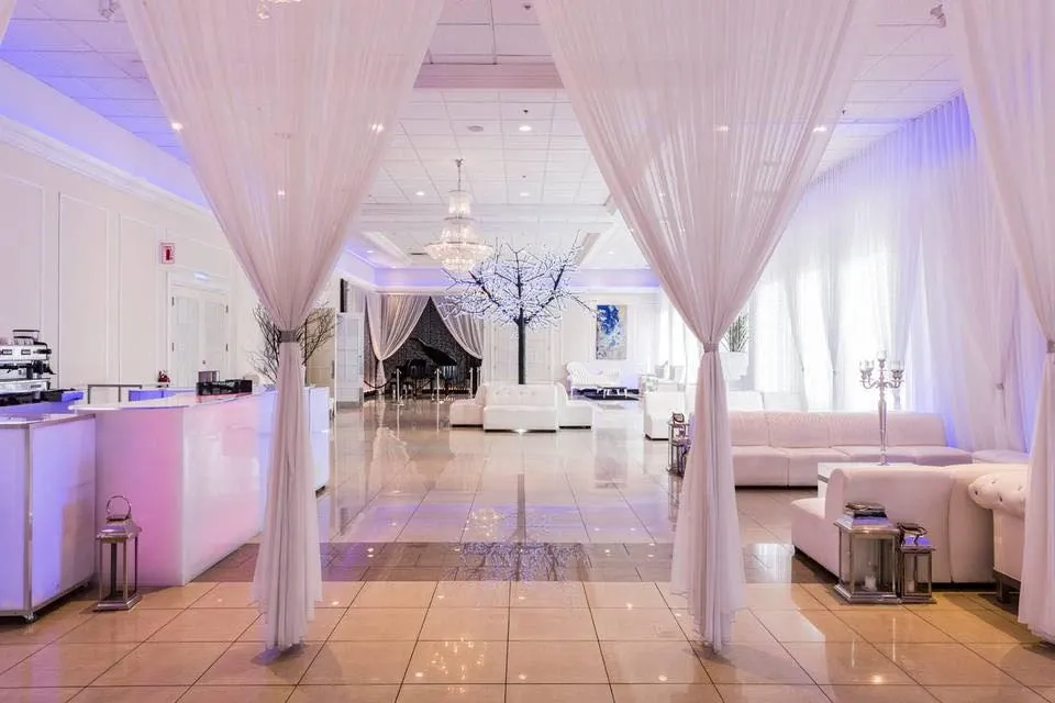 The image showcases the elegant interior of Embassy Plaza, a top-tier wedding venue listed on Mariage Elegant in Canada. The spacious room features a polished tiled floor and is adorned with flowing white drapes that create a soft, romantic ambiance. Crystal chandeliers hang from the ceiling, casting a gentle glow over the area. White sofas are tastefully arranged, offering a comfortable space for guests. Charming candelabras and delicate branches add a touch of sophistication to the decor, making it an ideal setting for memorable wedding celebrations.