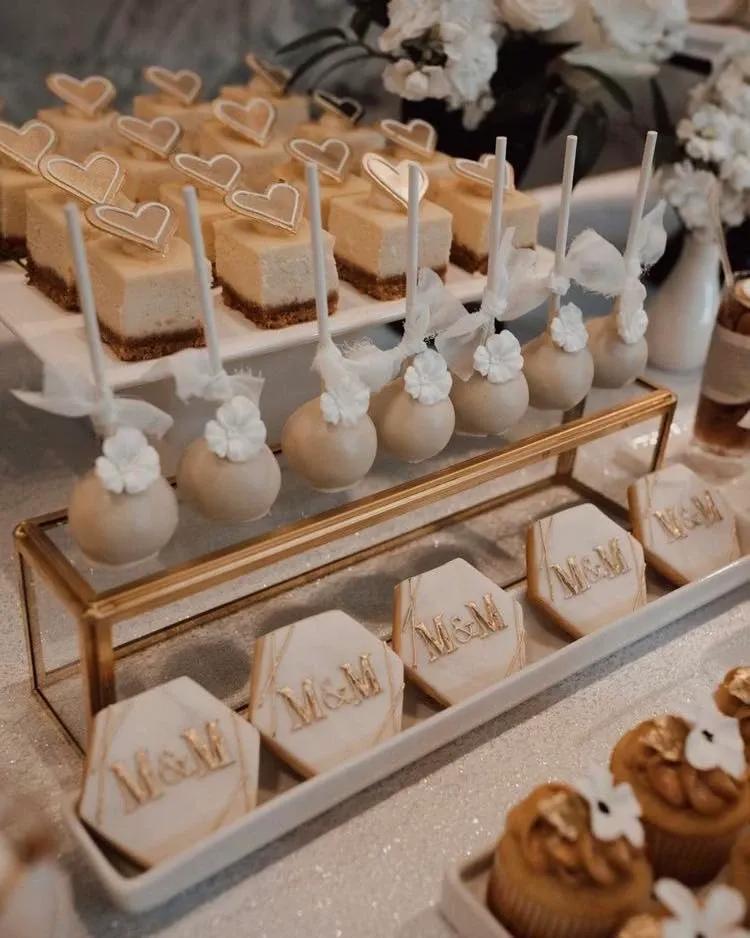 The image showcases a beautifully arranged dessert table at Embassy Plaza, a renowned wedding venue under the wedding venues category on Mariage Elegant, Canada. The table is elegantly decorated with a variety of sweets, including heart-shaped cheesecakes, cake pops adorned with delicate white flowers, and intricately designed cookies featuring the initials 'M & M.' The display also includes gold-accented cupcakes, contributing to the sophisticated and luxurious atmosphere expected at a wedding event. The desserts are carefully placed on gold and white stands, surrounded by lush white floral arrangements, adding to the overall aesthetic appeal for couples seeking an elegant setting for their special day.