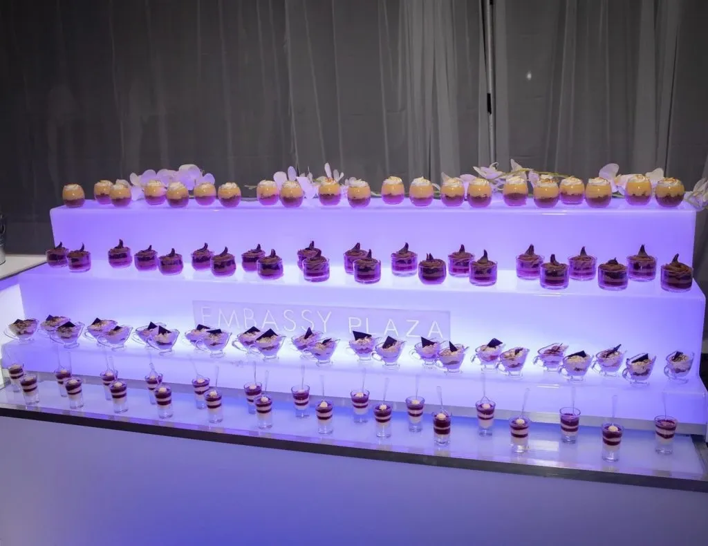 The image showcases a beautifully arranged dessert table at Embassy Plaza, a premier wedding venue in Canada. The table features three elegant tiers, each adorned with a variety of artfully crafted desserts, including cupcakes and layered parfaits in delicate glassware. The display is illuminated with a soft purple glow, enhancing the luxurious and inviting ambiance. The name 'Embassy Plaza' is subtly visible, reinforcing the venue's identity. This setting exemplifies the exquisite culinary presentation one can expect when choosing Embassy Plaza for a wedding celebration.