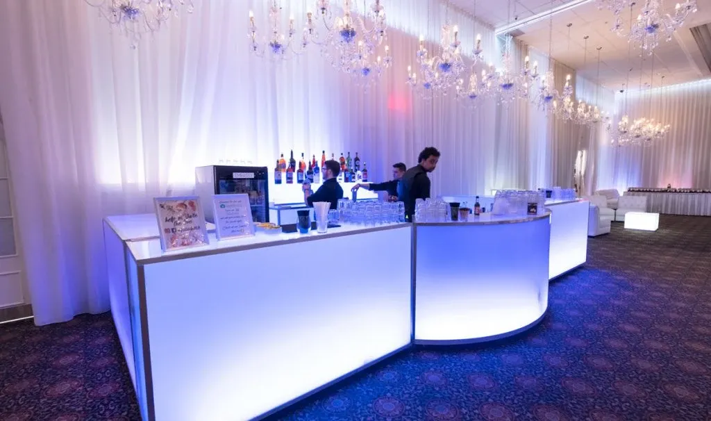 The image shows a reception area at Embassy Plaza, categorized as a wedding venue, which features a sophisticated bar setup. Positioned under opulent chandeliers, the illuminated bar counter casts a soft glow, enhancing the elegant ambiance ideal for weddings. Bartenders are seen preparing drinks, and an array of colorful bottles is neatly arranged in the background. The room is adorned with white drapery and subtle lighting, contributing to a luxurious and inviting atmosphere for wedding celebrations.