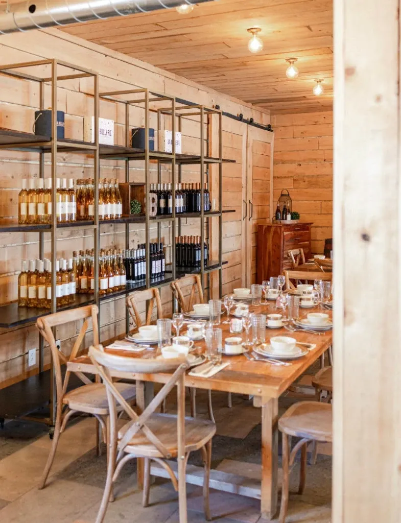 This image showcases the interior of La Bullerie, a wedding venue listed under Mariage Elegant in Canada. The rustic dining room features a long wooden table elegantly set with fine china, glassware, and dining utensils, reflecting a warm and intimate setting perfect for wedding receptions. The background highlights wooden shelves adorned with an assortment of wine bottles, adding a sense of sophistication and charm. The wooden walls and soft overhead lighting create a cozy and inviting atmosphere that complements the overall aesthetic of this unique wedding venue.