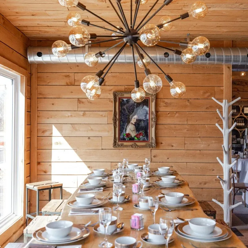 The image showcases a charming wedding venue by La Bullerie, categorized under Wedding Venues, featured on Mariage Elegant, Canada. It presents a well-decorated dining area with a long wooden table set for a gathering. The table is elegantly arranged with white china, glassware, and silver cutlery, creating a welcoming atmosphere. Above the table hangs a modern chandelier with multiple light bulbs, adding a contemporary touch to the rustic wooden interior. A framed painting adorns the wooden wall, enhancing the cozy and inviting ambiance suitable for wedding celebrations.