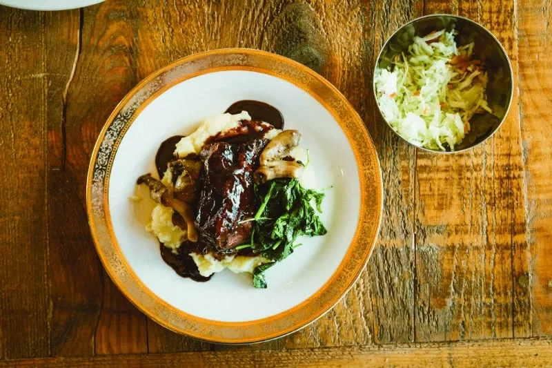 The image showcases a beautifully presented dish on a rustic wooden table at La Bullerie, a distinguished wedding venue in Canada under the Mariage Elegant listings. The dish features a rich, dark sauce drizzled over a generous portion of mashed potatoes, topped with sautéed mushrooms and a side of wilted greens. Accompanying this main dish is a small bowl of fresh coleslaw, adding a crisp texture and vibrant color to the setting. La Bullerie offers exquisite culinary experiences, making it an ideal choice for wedding celebrations.