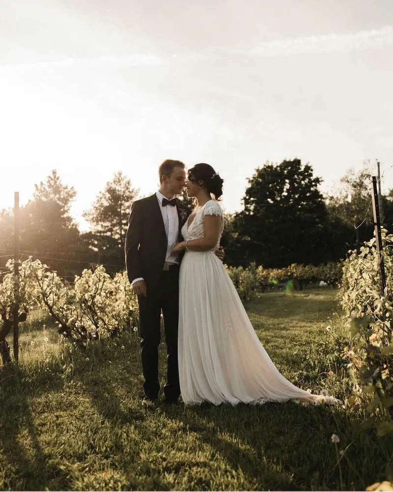 A couple stands closely together in an elegant vineyard setting at La Bullerie, a renowned wedding venue in Canada. Bathed in the warm glow of a setting sun, the bride wears a flowing white gown with lace details, and the groom is dressed in a classic black tuxedo. The lush green vineyard surrounds them, adding a romantic and serene ambiance, perfect for weddings. The sun casts a golden hue over the landscape, highlighting the natural beauty of the location. This image exemplifies the enchanting wedding experiences offered by La Bullerie.