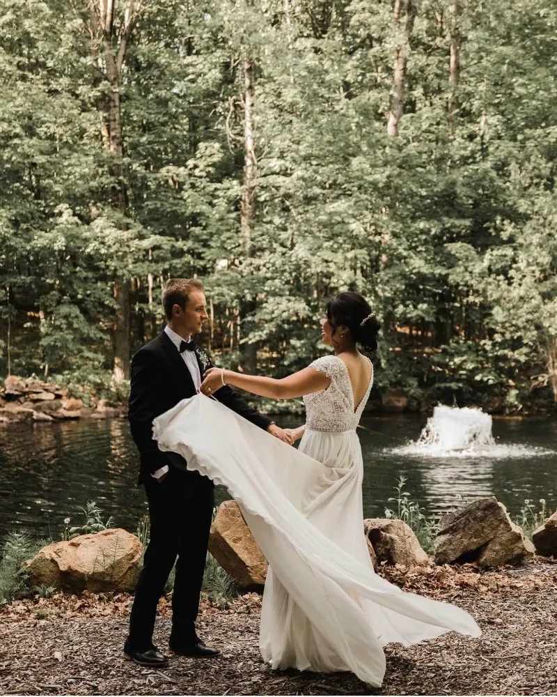 A bride and groom stand beside a serene pond surrounded by lush greenery at La Bullerie, a wedding venue in Canada. The groom is dressed in a classic black tuxedo, while the bride wears an elegant white gown with lace detailing, its long train gracefully spread out. In the background, a small fountain creates gentle ripples in the pond, adding to the romantic and tranquil setting. This image captures the scenic beauty prevalent at La Bullerie, making it an ideal choice for a memorable wedding venue.