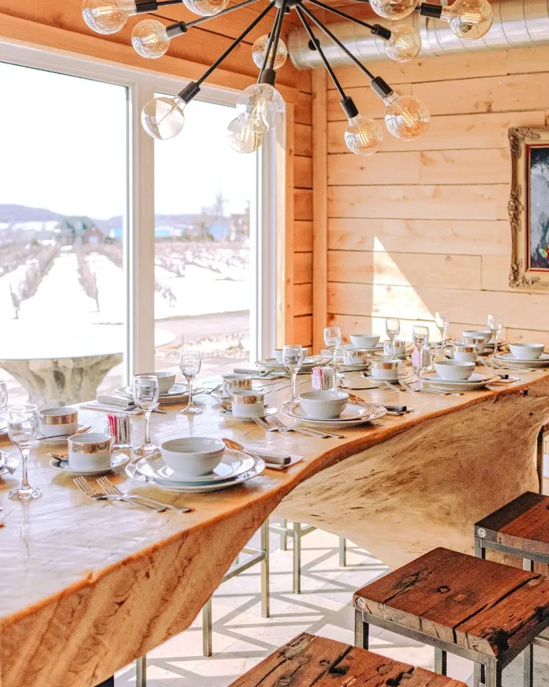 The image showcases a beautifully arranged wedding venue by La Bullerie, categorized under Wedding Venues on Mariage Elegant, Canada. The setting includes a long, rustic wooden table adorned with elegant table settings, including white china, glassware, and silverware. The room is well-lit by large, contemporary light fixtures and natural light streaming in through the large window, which offers a view of a scenic vineyard and snow-covered landscape beyond. Wooden benches provide seating, accentuating the rustic charm of the space. The ambiance is warm and inviting, perfect for an intimate wedding gathering.