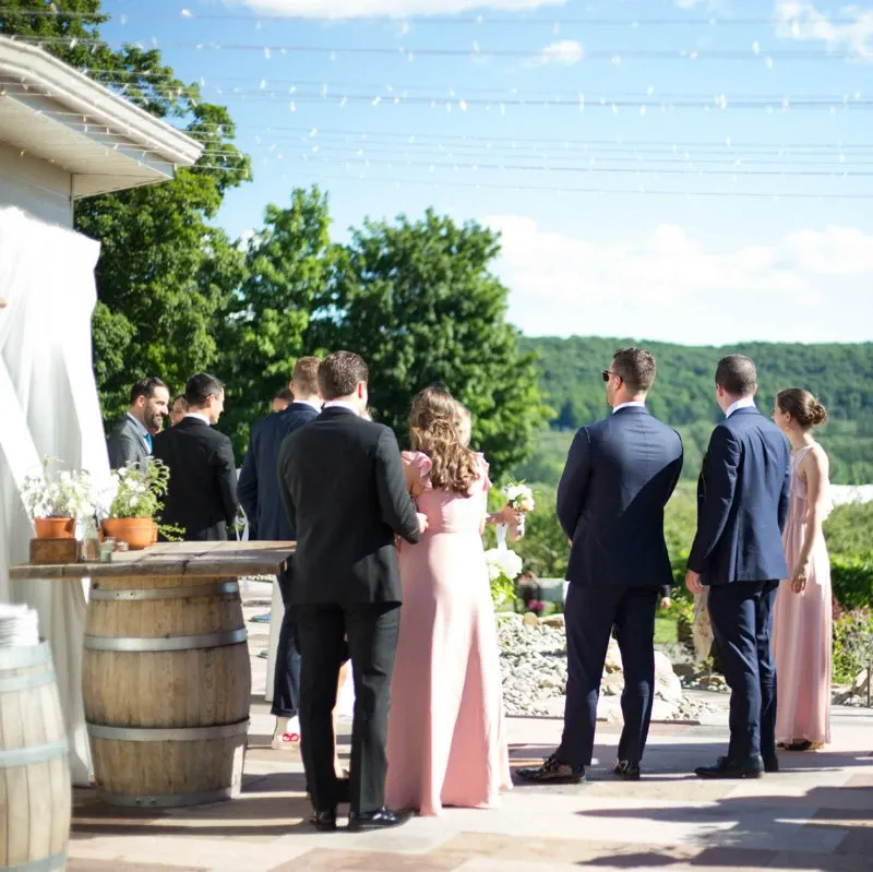 In this image, a group of elegantly dressed wedding guests, including men in suits and women in pastel pink dresses, are gathered outdoors at the La Bullerie wedding venue. The setting features lush greenery and a scenic view of the surrounding hills, providing a picturesque backdrop. Overhead string lights suggest a charming and festive atmosphere, ideal for a wedding celebration. Wooden barrels serve as decorative elements, enhancing the rustic charm of the venue. La Bullerie is highlighted here as a beautiful option for couples seeking an enchanting wedding venue in Canada.