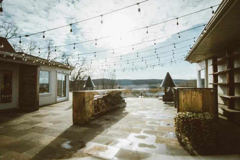 The image shows an expansive outdoor terrace at La Bullerie, a wedding venue in Canada listed on Mariage Elegant. String lights are elegantly draped overhead, enhancing the romantic ambiance of the setting. The terrace features a stone floor and is surrounded by rustic wooden structures, providing an ideal space for wedding receptions or events. Beyond the terrace, scenic views of rolling hills and a clear sky create a picturesque backdrop, emphasizing the venue's natural beauty.