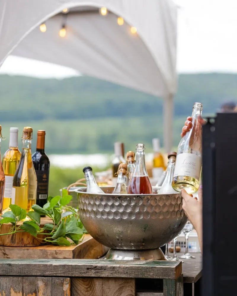 The image showcases an elegant outdoor bar setup at La Bullerie, a wedding venue listed on Mariage Elegant in Canada. A variety of wine bottles, some open and some sealed, are displayed in a large, textured metal ice bucket on a rustic wooden counter. A hand is seen reaching for a bottle labeled 'Kramer.' The area is adorned with green leafy decorations and wooden crates, creating a charming rustic atmosphere. In the background, a white canopy is visible, suggesting preparedness for an outdoor wedding event, with scenic green hills and a water body further enhancing the picturesque setting.