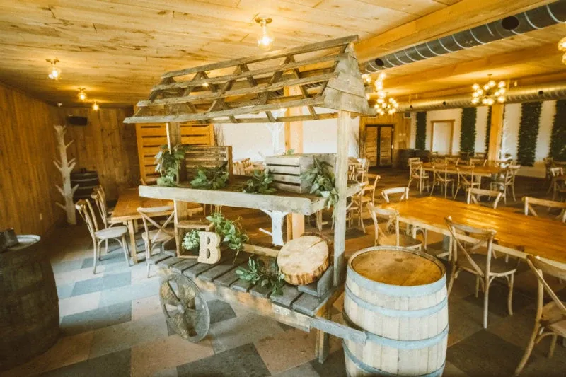 The image showcases La Bullerie, a charming wedding venue under the category of Wedding Venues on Mariage Elegant, Canada. The rustic interior features wooden decor and warm lighting, creating an inviting atmosphere. There is a decorative wooden cart adorned with greenery, wooden crates, and a cut log, adding to the rustic charm. Surrounding the cart are wooden tables and chairs neatly arranged for a reception. The walls are lined with vertical wooden panels and greenery accents, contributing to the venue's natural and cozy ambiance.
