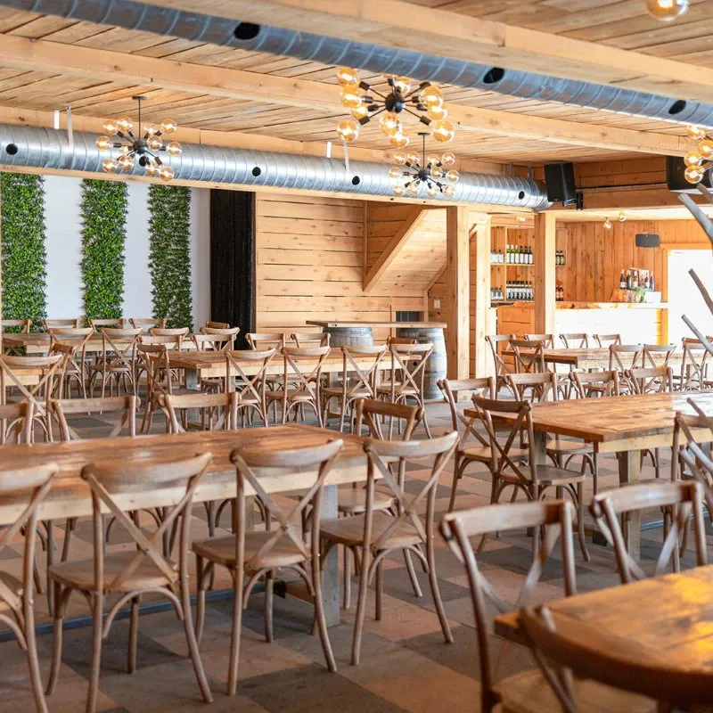 The image showcases the interior of La Bullerie, a wedding venue listed under the wedding venues category on Mariage Elegant, Canada. The venue features a rustic and elegant setting with wooden tables and chairs arranged neatly across the space. The ceiling is adorned with industrial-style exposed ducts and unique light fixtures, adding a modern touch. Greenery is displayed along one wall, contributing to a natural and inviting atmosphere. A wooden bar area is visible in the background, enhancing the venue's charm and readiness to host wedding receptions.