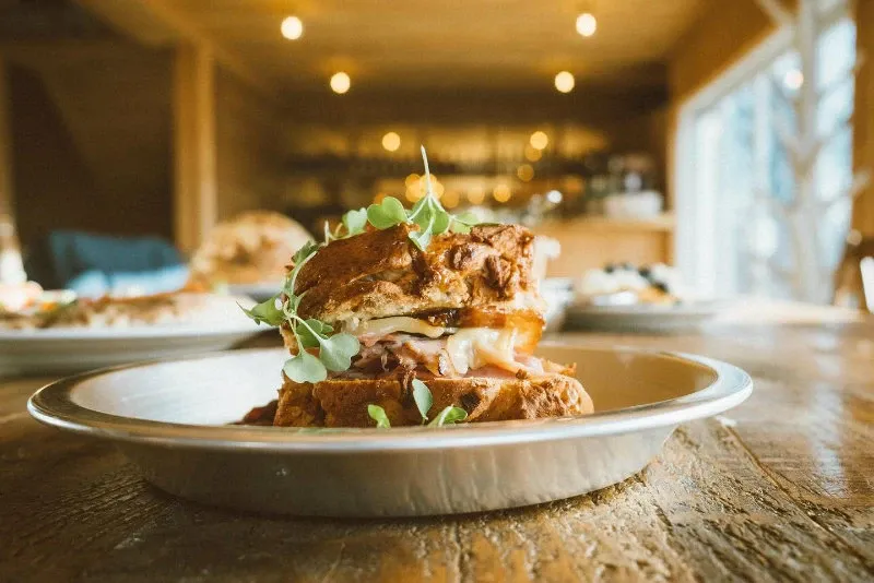 The image showcases a beautifully plated gourmet sandwich on a rustic wooden table, situated within the elegant setting of La Bullerie, a wedding venue in Canada. Natural light floods through a large window, creating a warm and inviting atmosphere, ideal for a wedding reception. The sandwich, artfully garnished with fresh greens, hints at the exquisite catering options available for celebrations at this venue.