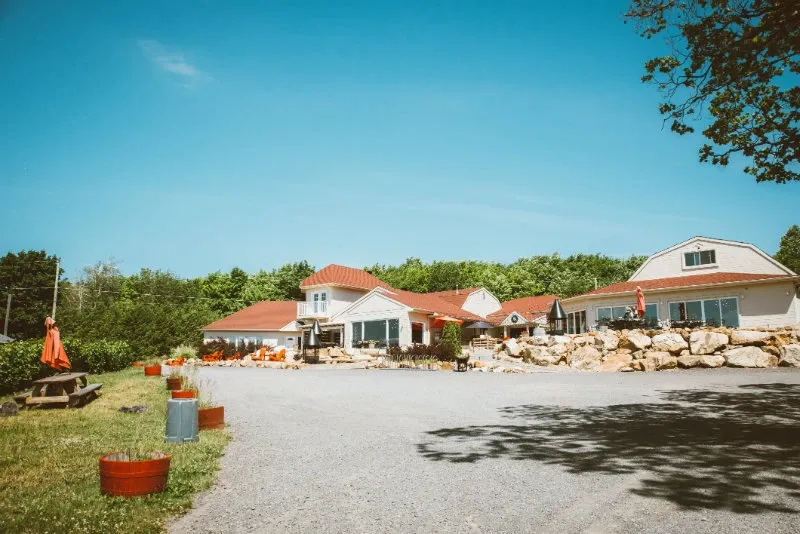 La Bullerie, a stunning wedding venue in Canada, is showcased in this image. The venue features a charming, rustic building with red rooftops surrounded by lush greenery. A spacious gravel driveway leads to the entrance, with well-maintained landscaping and decorative stone accents adding to the picturesque setting. Bright patio umbrellas and outdoor seating suggest a welcoming space for guests, perfect for a memorable wedding celebration.