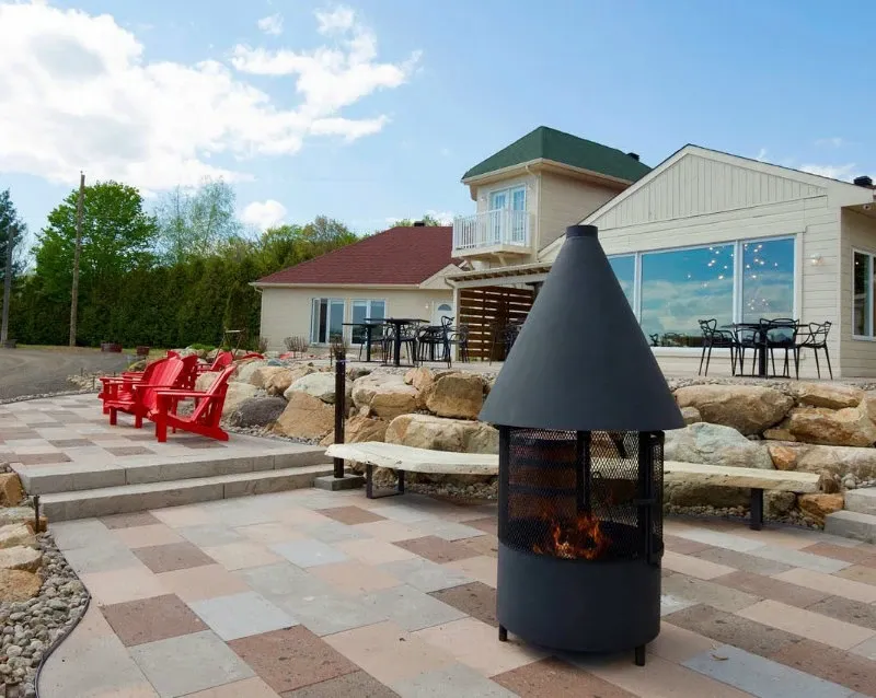 The image showcases La Bullerie, a wedding venue in Canada listed on Mariage Elegant. The picturesque scene includes a modern building with large windows, set in a tranquil outdoor area. In the foreground, there is a stylish outdoor fireplace surrounded by a neatly tiled patio. A series of red Adirondack chairs are positioned on the left, facing a serene landscape, ideal for wedding guests to relax. The venue's modern architectural design blends with the natural surroundings, providing a beautiful setting for weddings and receptions. The backdrop of trees and an open sky adds to the charm and appeal of La Bullerie as an exquisite location for ceremonies.