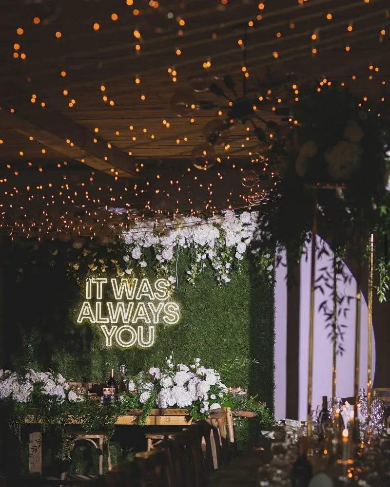 The image showcases a beautifully decorated wedding venue by La Bullerie, categorized under Wedding Venues on Mariage Elegant, Canada. The venue features an elegant setting with a lush green backdrop adorned with white floral arrangements. A prominent neon sign reading 'It was always you' adds a romantic touch to the ambiance. Warm, ambient string lights hang from the ceiling, creating a cozy and inviting atmosphere perfect for a wedding celebration. The table settings are elegantly arranged with floral accents, enhancing the venue's sophisticated and festive feel.