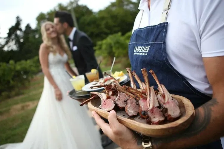 In this image of La Bullerie, a notable wedding venue in Canada, a server in a branded apron elegantly presents a platter of gourmet lamb chops alongside a selection of appetizers. In the background, a newlywed couple shares a tender moment, with the bride in a flowing white gown and the groom in a dark suit, set amidst the lush greenery of a vineyard, highlighting the romantic and picturesque setting ideal for weddings.