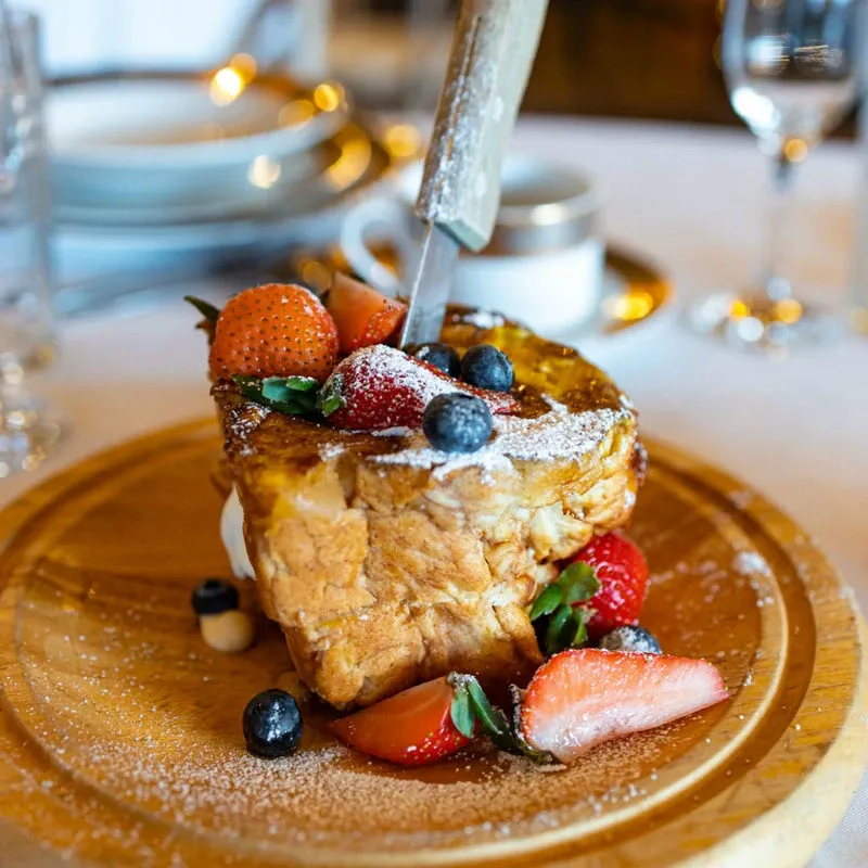 This image showcases a beautifully presented dessert at La Bullerie, a wedding venue featured in the Mariage Elegant vendor listings in Canada. A thick slice of brioche toast is artistically garnished with fresh strawberries, blueberries, and a dusting of powdered sugar. The dessert is placed on a wooden platter, with elegant glassware and stacked plates subtly visible in the background, highlighting the refined dining experience available at this wedding venue.