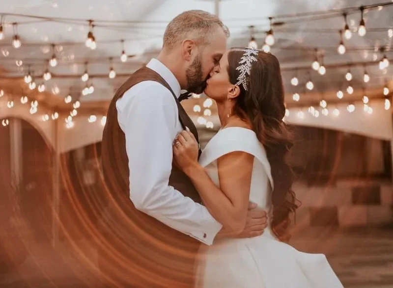 A newlywed couple shares a kiss at La Bullerie, a premier wedding venue in Canada featured on Mariage Elegant. The groom is dressed in a formal vest and bow tie, while the bride wears an elegant off-shoulder gown with a sparkling headpiece. They are surrounded by ambient string lights, creating a romantic atmosphere. The gentle swirl in the foreground adds a dreamy effect to this intimate moment, showcasing the enchanting setting of La Bullerie for weddings.