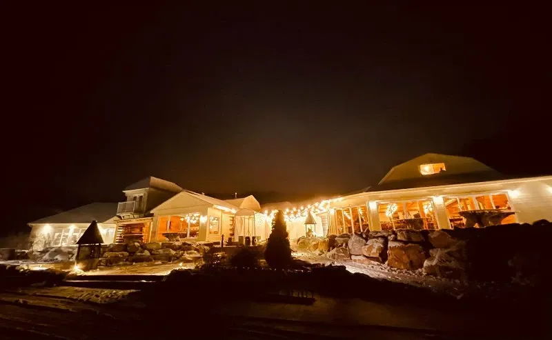 La Bullerie, listed under the wedding venues category, is beautifully illuminated at night in this image. The venue, showcasing elegant architecture, features cozy, warmly lit interiors visible through large windows. String lights adorn the exterior, creating a romantic and inviting atmosphere ideal for weddings. The surrounding landscape is softly lit, highlighting natural stone features that add to the charm of the location. This picturesque venue in Canada is perfect for couples seeking a magical wedding setting.