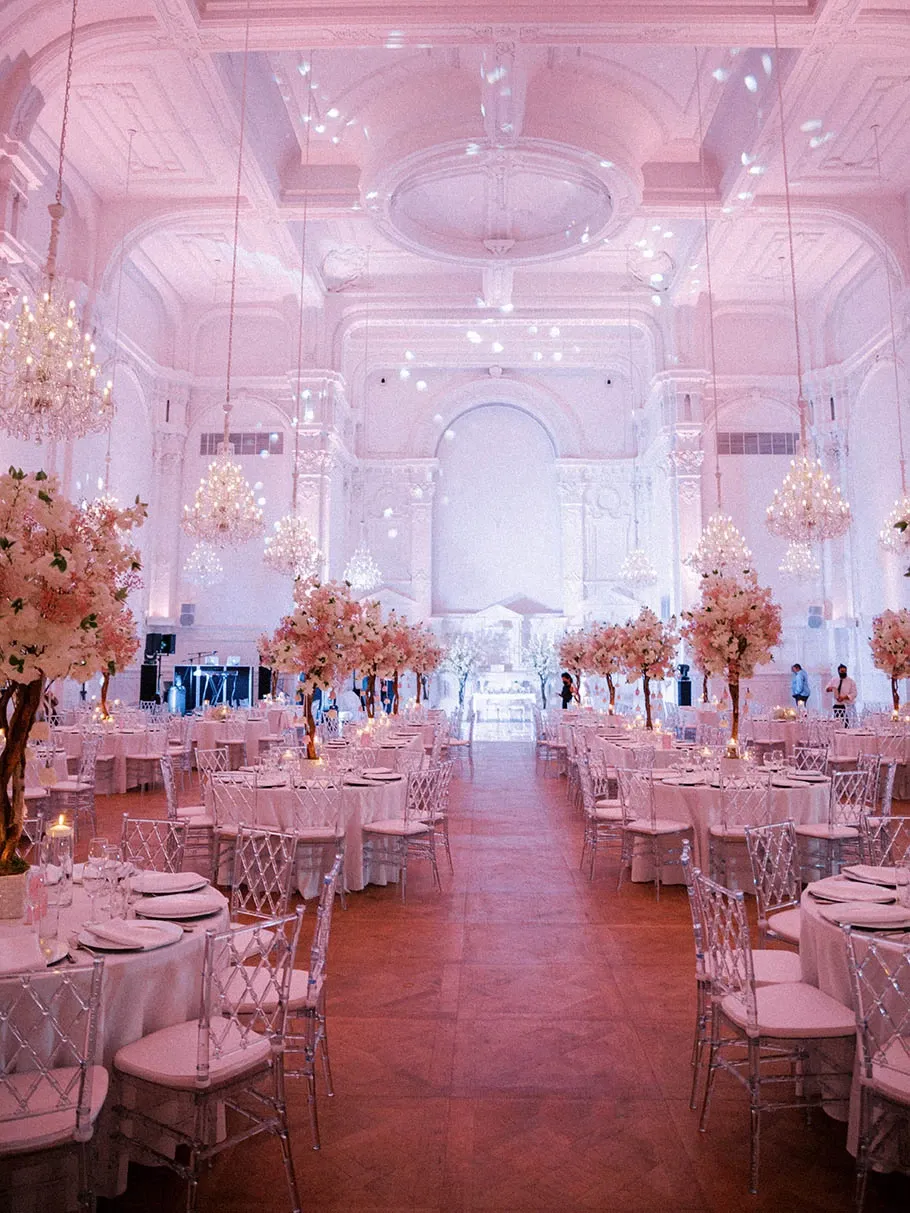 The image showcases a grand wedding venue by Maison Principale, categorized under Wedding Venues, featured on Mariage Elegant in Canada. The venue is elegantly decorated with high ceilings and opulent chandeliers, casting a warm glow across the room. The tables are adorned with white tablecloths and set with fine dinnerware, surrounded by transparent, intricately designed chairs. Lush floral arrangements featuring pink and white blossoms are placed atop tall, golden stands, lining the central aisle. The lavish setting is complemented by a beautifully lit backdrop, creating a romantic and sophisticated atmosphere ideal for a wedding celebration.