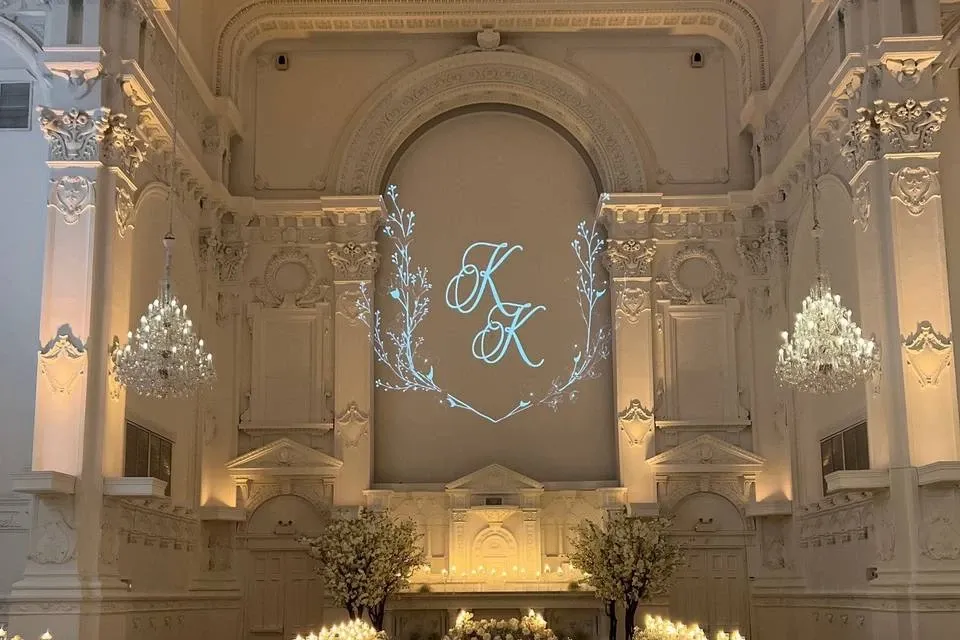 The image showcases the interior of Maison Principale, a premier wedding venue listed on Mariage Elegant in Canada. The grand hall features intricate architectural details with high, ornate ceilings and elegant moldings. Two large chandeliers hang prominently, casting a soft glow over the space, which highlights the elegance of the decor. In the center, a beautifully arranged altar is adorned with lush white floral arrangements, framed by delicate lighting. Above the altar, the initials 'K & K' are projected in an elegant script, adding a personal touch to the sophisticated ambiance, ideal for creating memorable wedding ceremonies.