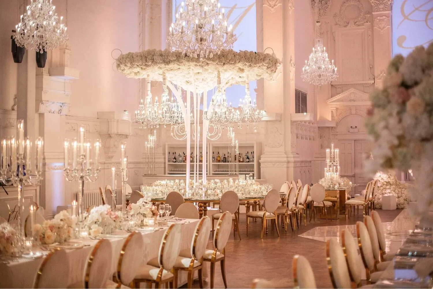 The image showcases an opulent wedding venue by Maison Principale, under the Wedding Venues category, featured on Mariage Elegant, Canada. The elegant ballroom is adorned with lavish crystal chandeliers hanging from a high, beautifully decorated ceiling. Luxurious floral arrangements with white and blush flowers are centrally placed on towering stands, adding grandeur to the setting. The tables are elegantly set with fine china and crystal glassware, surrounded by plush chairs with gold accents. The ambience is further enhanced by the soft, romantic lighting, creating a perfect backdrop for a sophisticated wedding celebration.