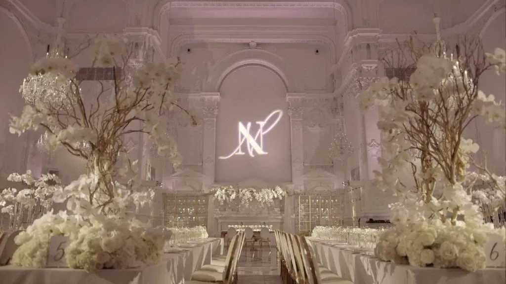 The image showcases an elegant wedding reception venue set up by Maison Principale, categorized under Wedding Venues. The spacious hall features high ceilings adorned with intricate architectural details and soft lighting, creating a romantic ambiance. The tables are beautifully arranged with white floral centerpieces and gold accents, while crystal chandeliers add a touch of luxury. In the background, the initials 'MP' are illuminated, highlighting the venue's sophisticated branding. This setting exemplifies a blend of classic and contemporary styles, perfect for a memorable wedding celebration.