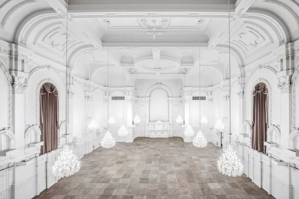 The image shows a stunning ballroom within Maison Principale, a premier wedding venue in the wedding planning and vendor listing site, Mariage Elegant, Canada. The elegant space features magnificent white walls adorned with intricate moldings and high, ornate ceilings. A series of grand chandeliers hang from above, casting a soft, inviting glow over the spacious hardwood floor, perfect for receptions and dance events. Rich, dark drapes frame the tall windows along the walls, adding a touch of luxury and sophistication to the venue. Maison Principale offers an exquisite setting for weddings, combining classical architecture with timeless elegance.