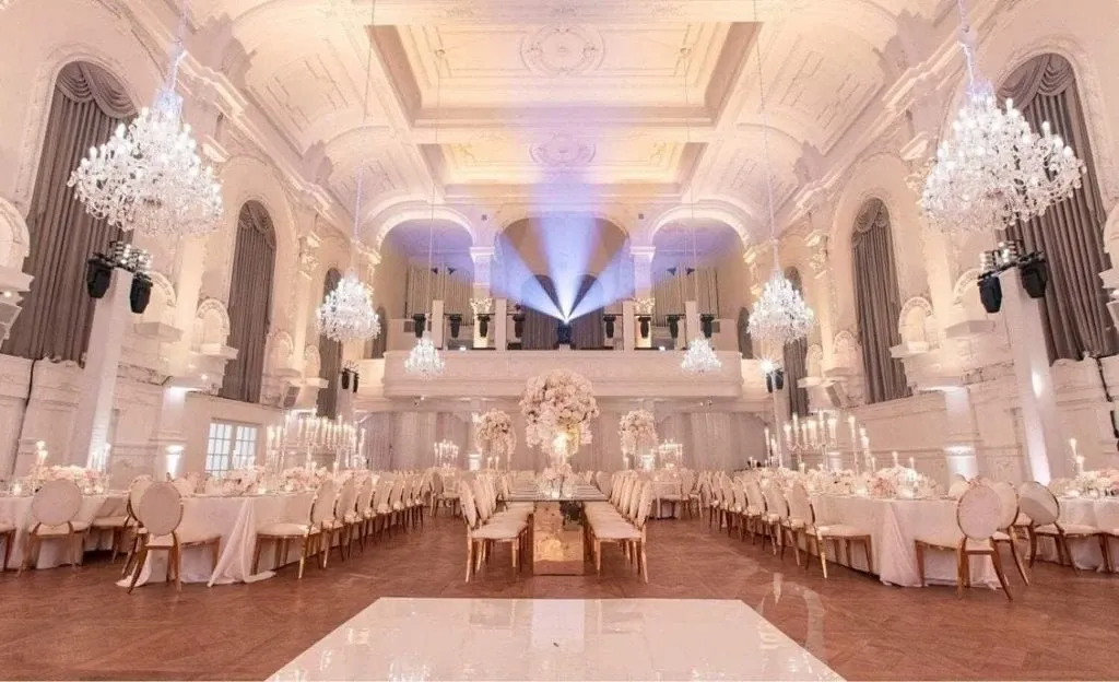 In this image of the Maison Principale, a premier wedding venue listed on Mariage Elegant, Canada, the opulent interior showcases a grand ballroom exquisitely decorated for a wedding reception. The high ceilings are adorned with multiple shimmering chandeliers casting a warm and inviting glow over the elegantly set tables. Each table is meticulously arranged with ivory linens, gold-rimmed chairs, and tall floral centerpieces featuring soft pink and white blooms. The vast room features ornate architectural details, including arched windows and intricate moldings, adding to the majestic ambiance suitable for a luxurious wedding event.