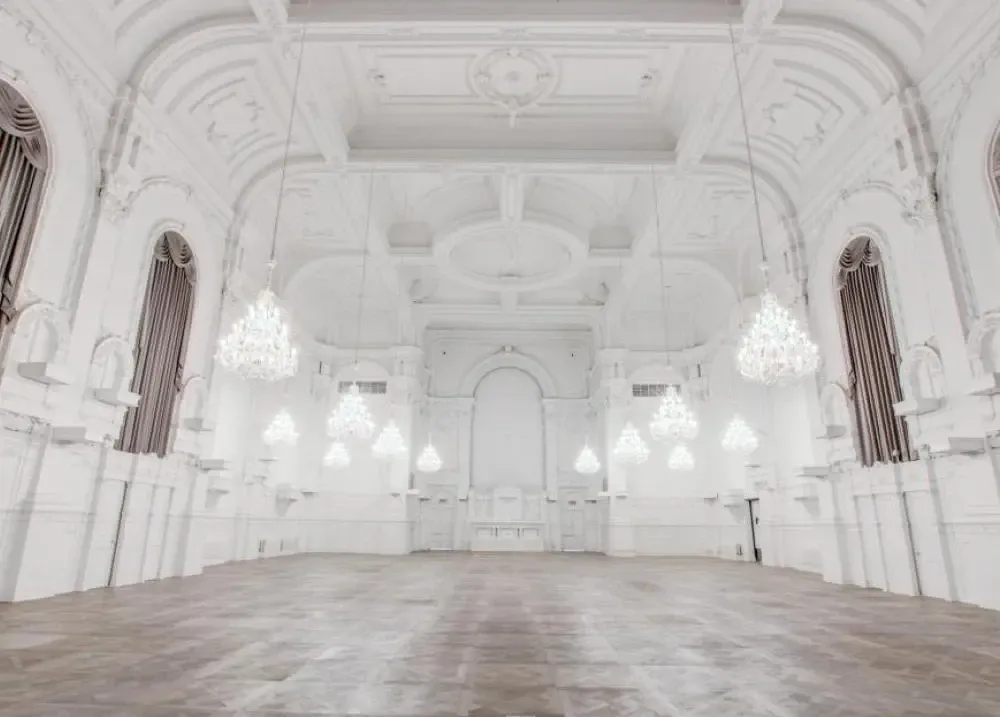 The image shows a grand ballroom at Maison Principale, a distinguished wedding venue listed on Mariage Elegant in Canada. The room features a spacious, elegant interior with high, ornate ceilings and intricate moldings. Multiple crystal chandeliers hang from above, casting a soft, luxurious glow across the polished wooden floor. Tall, arched windows with rich draperies add to the opulent setting, making it an ideal location for hosting sophisticated wedding ceremonies and receptions.