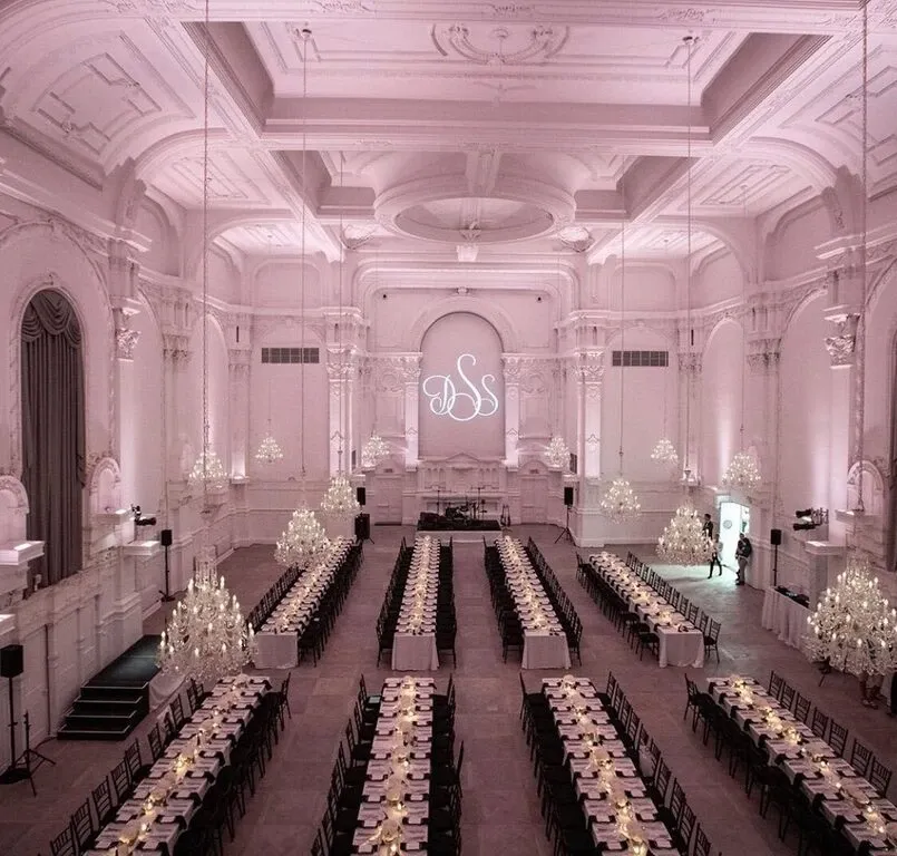 The image displays an opulent wedding venue at Maison Principale, categorized under Wedding Venues, featured on Mariage Elegant, Canada. The grand hall showcases an elegant setup with long dining tables adorned with white tablecloths and arranged with matching chairs. Each table is meticulously set with fine dinnerware and centerpieces. The spacious room is illuminated by numerous ornate chandeliers hanging from a beautifully detailed, high ceiling, creating a romantic and luxurious atmosphere. The soft pink and white lighting complements the sophisticated decor, highlighting the intricate architectural features and the initials projected above the stage area at the back. This setting embodies a perfect blend of elegance and grandeur, ideal for hosting a memorable wedding reception.
